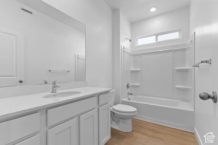 White vanity with countertop and integrated sink, polished chrome faucet, full-width mirror, tub-shower combination with integrated shelving, and wood-finish flooring