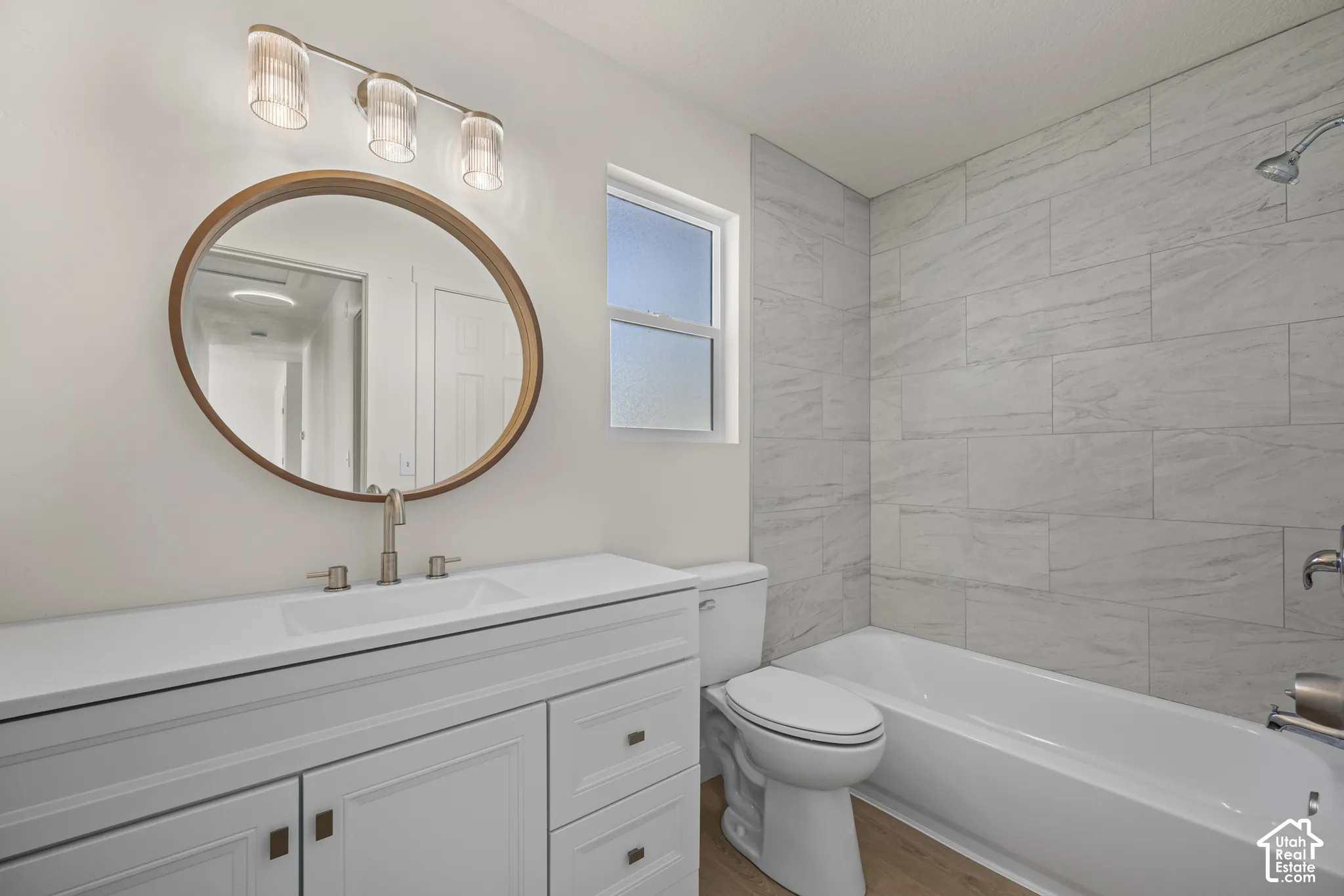 Bathroom featuring a white vanity with an integrated sink, round wood-framed mirror, three-light fixture, tub-shower combination with floor-to-ceiling tile surround, and wood-finish flooring