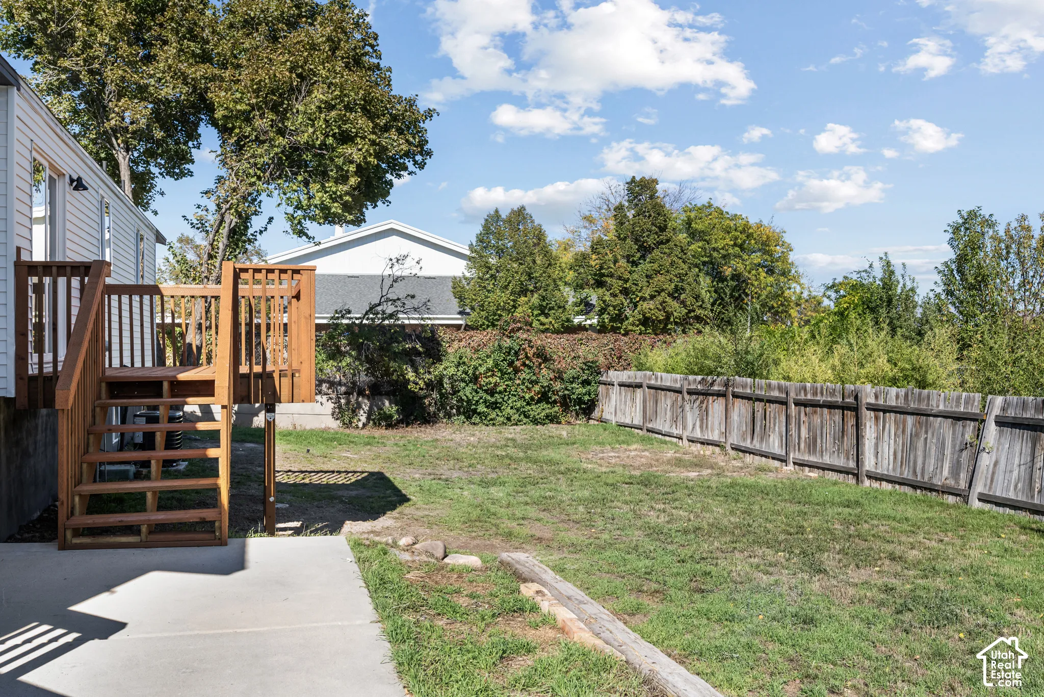 Backyard featuring a wood-tone raised deck with staircase, concrete patio, grass lawn, and wood privacy fencing