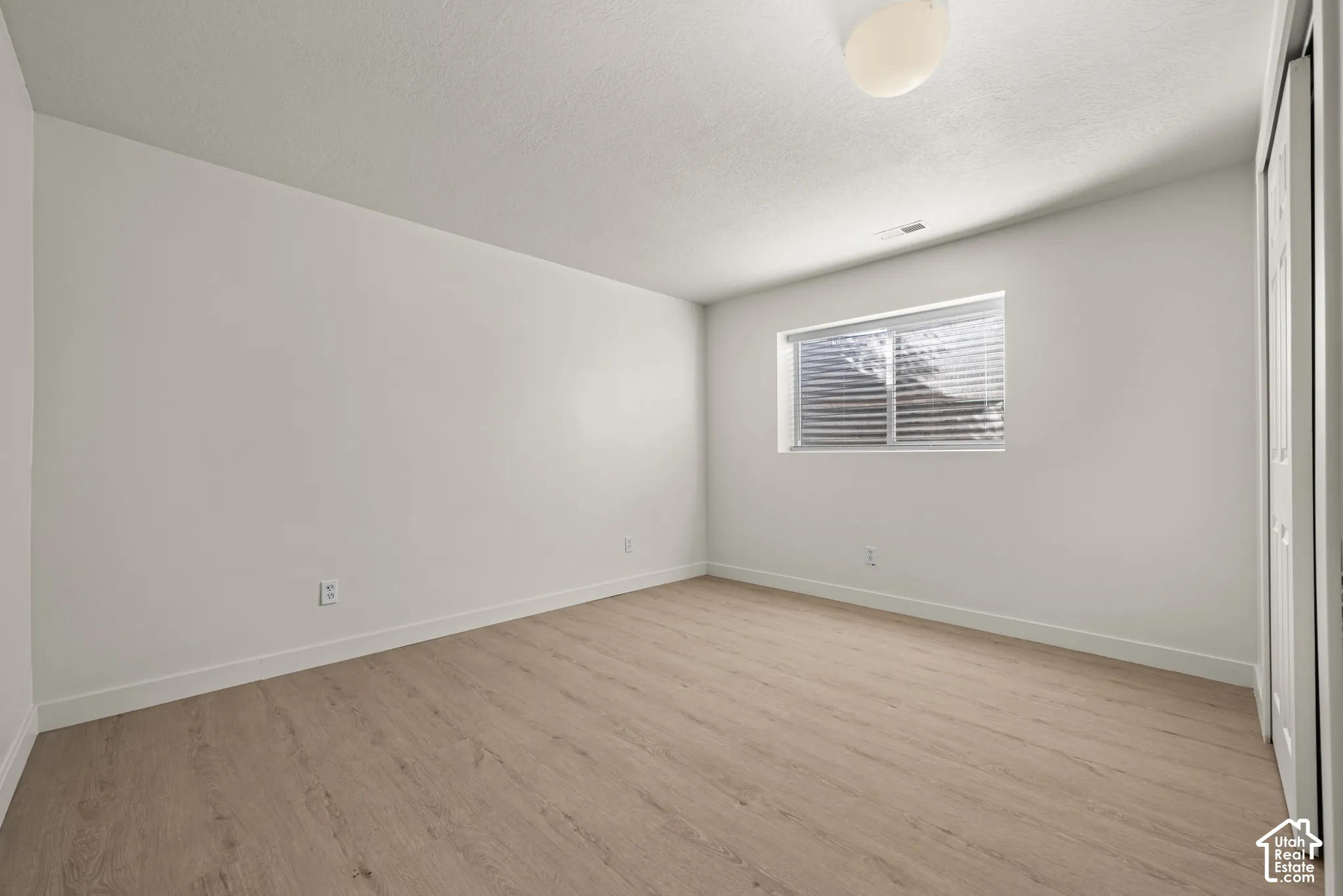 Neutral-toned room featuring wood-finish flooring, a single window, a ceiling-mounted light fixture, and a sliding-panel closet door