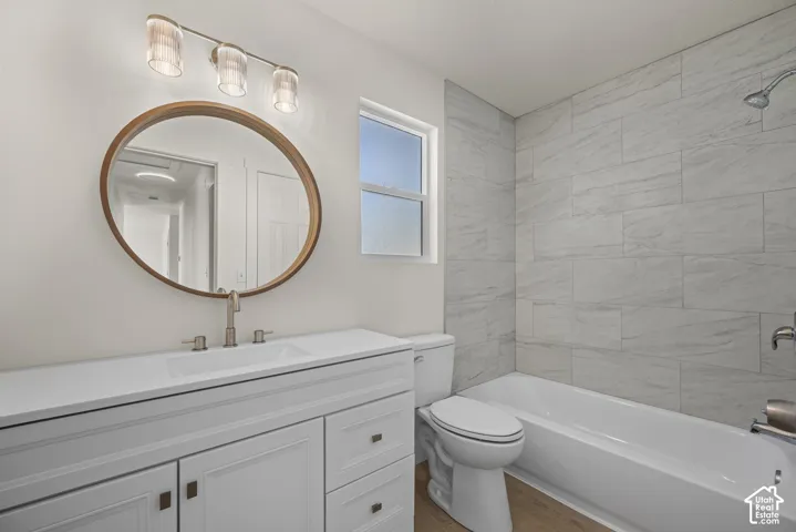 Bathroom featuring a white vanity with an integrated sink, round wood-framed mirror, three-light fixture, tub-shower combination with floor-to-ceiling tile surround, and wood-finish flooring