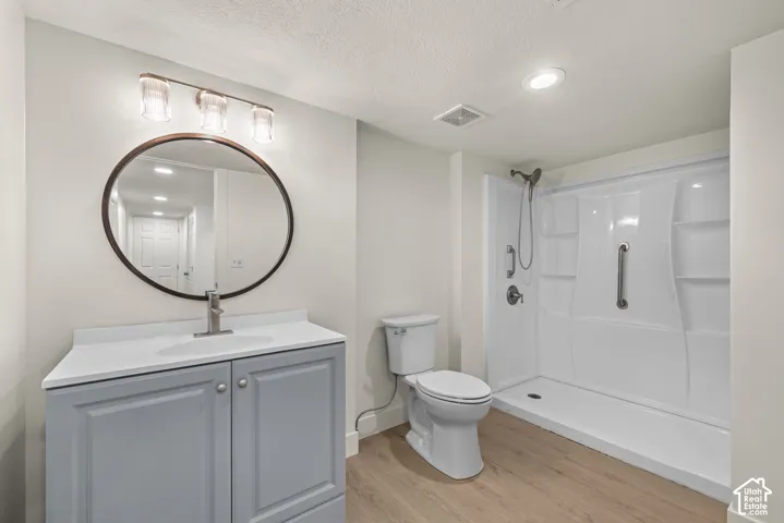 Bathroom featuring a gray vanity with white countertop, circular mirror, three-light fixture, and walk-in shower with integrated surround and wood-finish flooring