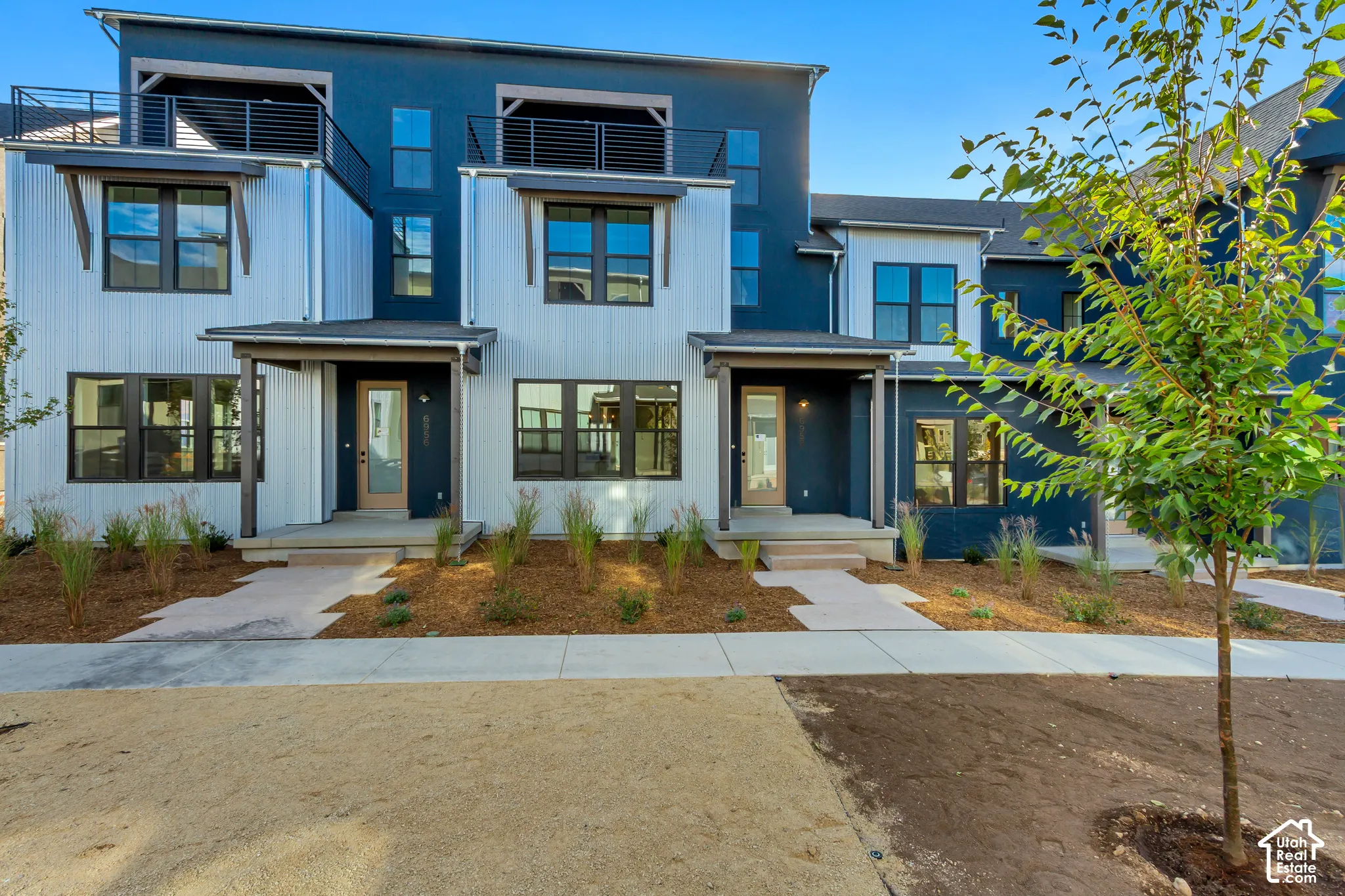 Modern multi-level exterior featuring a combination of vertical metal siding and dark-hued board and batten, upper-level balconies with black metal railings, and concrete entry porches