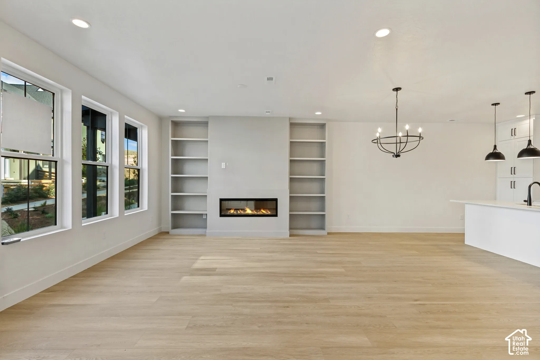 Open-concept area featuring light wood-finish flooring, a linear gas fireplace flanked by built-in shelving, expansive windows, recessed lighting, and a chandelier with multiple light sources