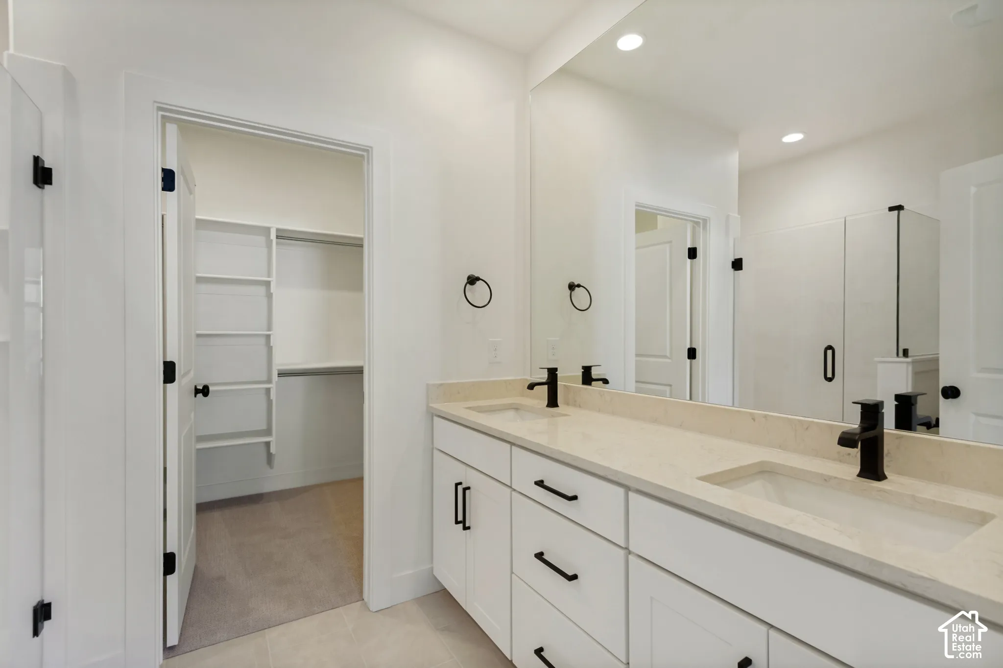 Double vanity with light-toned solid surface countertops, undermount sinks, matte black fixtures, white shaker-style cabinetry, and a full-width mirror