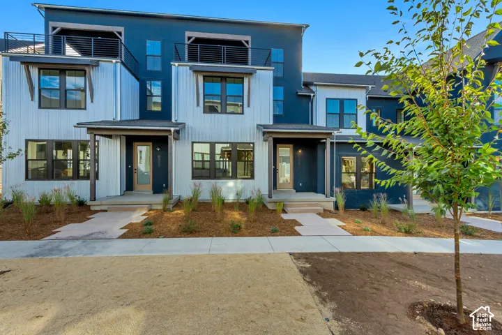 Modern multi-level exterior featuring a combination of vertical metal siding and dark-hued board and batten, upper-level balconies with black metal railings, and concrete entry porches
