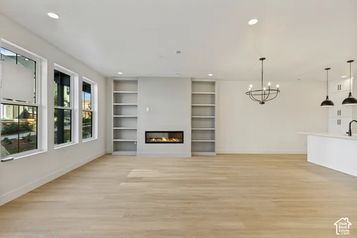 Open-concept area featuring light wood-finish flooring, a linear gas fireplace flanked by built-in shelving, expansive windows, recessed lighting, and a chandelier with multiple light sources