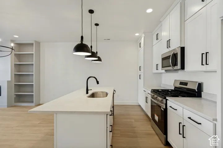 Kitchen featuring white cabinetry, an island with a stainless steel sink and high-arc faucet, stone countertops, stainless steel appliances, and wood-finish flooring with built-in shelving