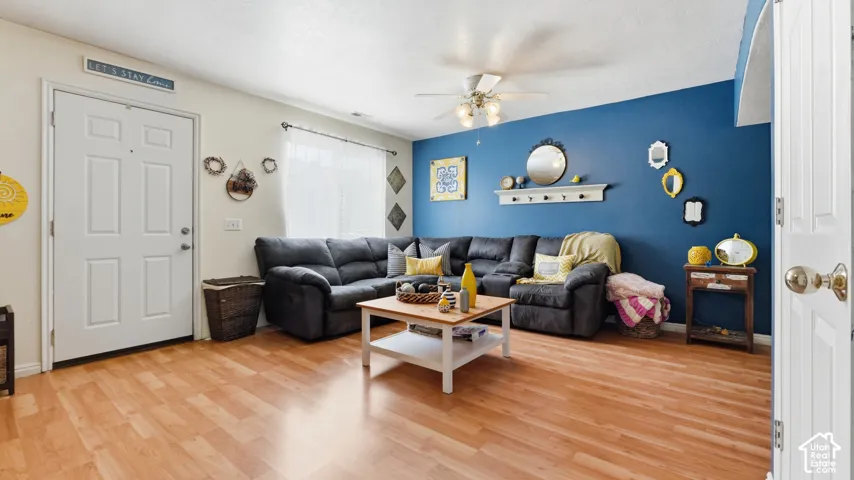 Living area featuring wood-finish flooring, a wall-mounted floating display shelf, and a ceiling fan with integrated lighting