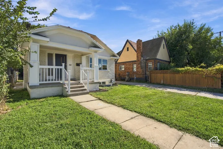 Light-colored brick exterior featuring a covered front porch, white railings, a concrete walkway, and a spacious front lawn