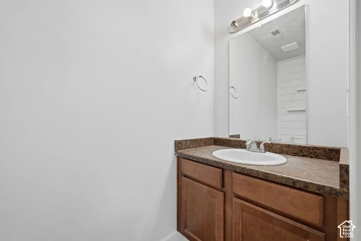Wood-finish cabinetry featuring an integrated vanity, stone-finish countertop with an oval drop-in sink, wall-mounted mirror, and three-light vanity fixture