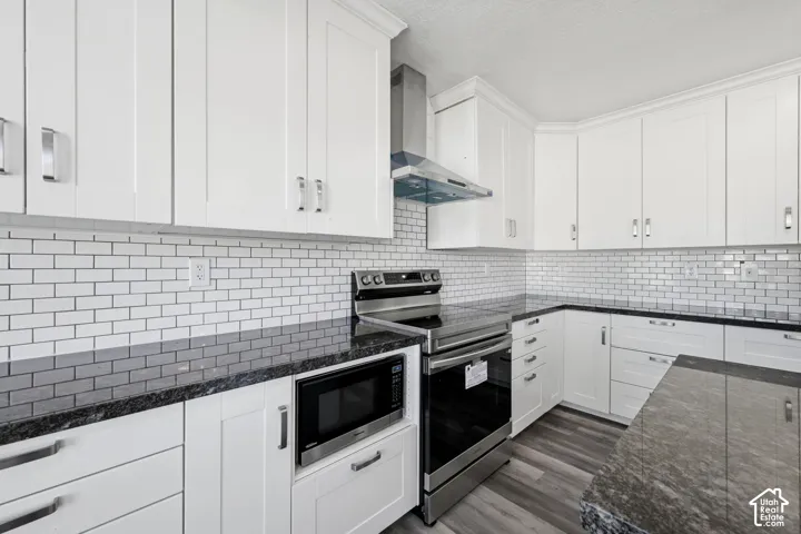 Kitchen featuring white shaker-style cabinetry, stone-finish countertops, subway tile backsplash, stainless steel range with overhead vent hood, and wood-finish flooring