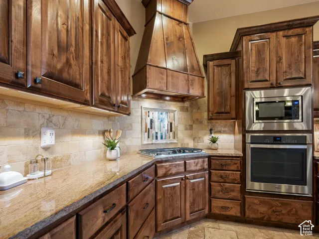 Custom stained distressed alder wood cabinetry, stone tile backsplash, built-in vent hood, stone countertops, and stainless steel wall-mounted oven and microwave combination