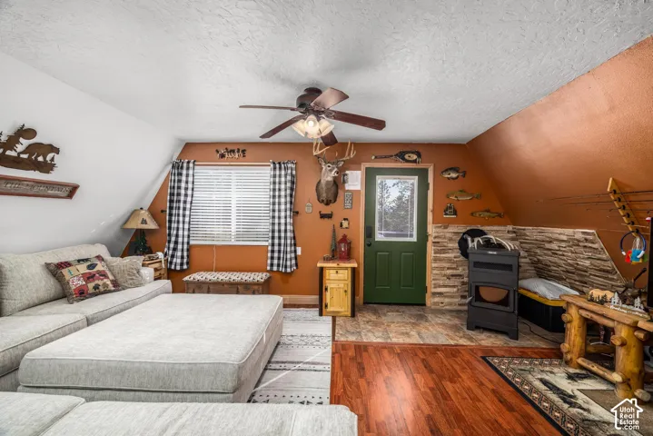 Sloped ceiling architecture featuring wood-finish flooring, a stone-accented wall, a metal stove, and a ceiling fan with integrated lighting