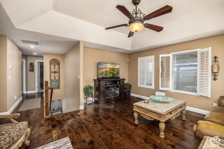 Tray ceiling with a central multi-blade ceiling fan, rich wood-finish flooring, and white plantation shutters