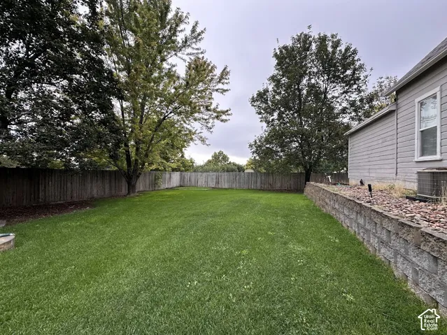 Backyard featuring a grass lawn, perimeter wood fencing, mature trees, and a tiered stone retaining wall