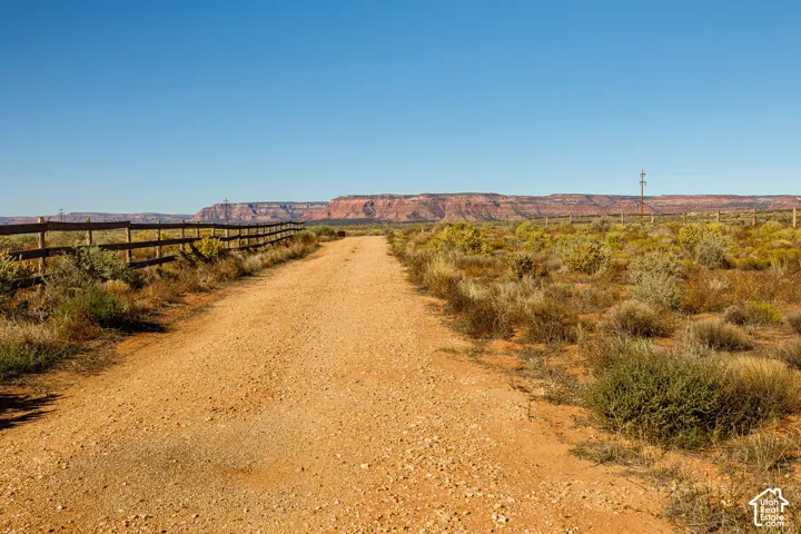 Expansive property featuring native desert vegetation, a gravel road, and a wooden post-and-rail fence set against a prominent red rock mesa backdrop