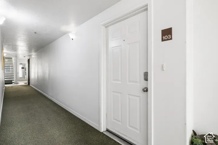 Hallway featuring neutral-toned carpeting, white walls, wall-mounted light fixtures, and a six-panel door with metal hardware