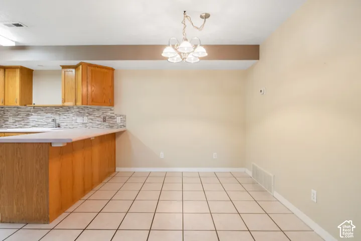 Kitchen area featuring wood cabinetry, a tiled backsplash, a peninsula with a solid-surface countertop, tile flooring, and a multi-light chandelier