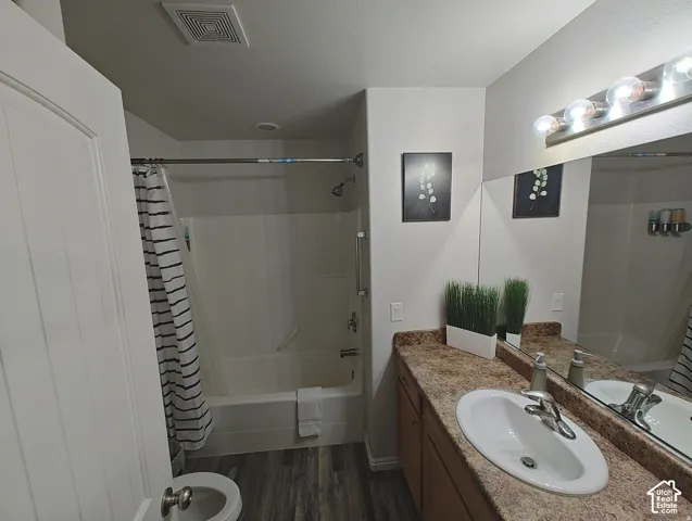 Bathroom featuring a vanity with a stone-finish countertop, integrated oval basin, full-width vanity mirror, multi-bulb overhead lighting, and a combination tub-and-shower enclosure with wood-finish flooring