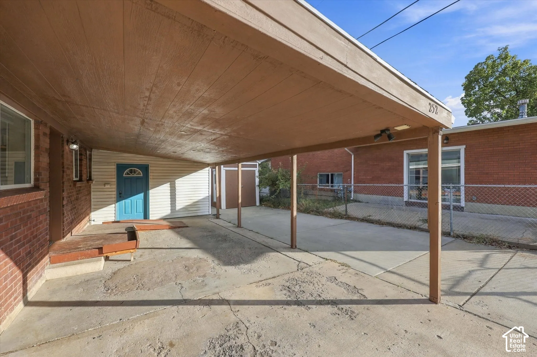 Covered carport with wood-finish ceiling, brick exterior siding, concrete driveway, and chain-link fencing