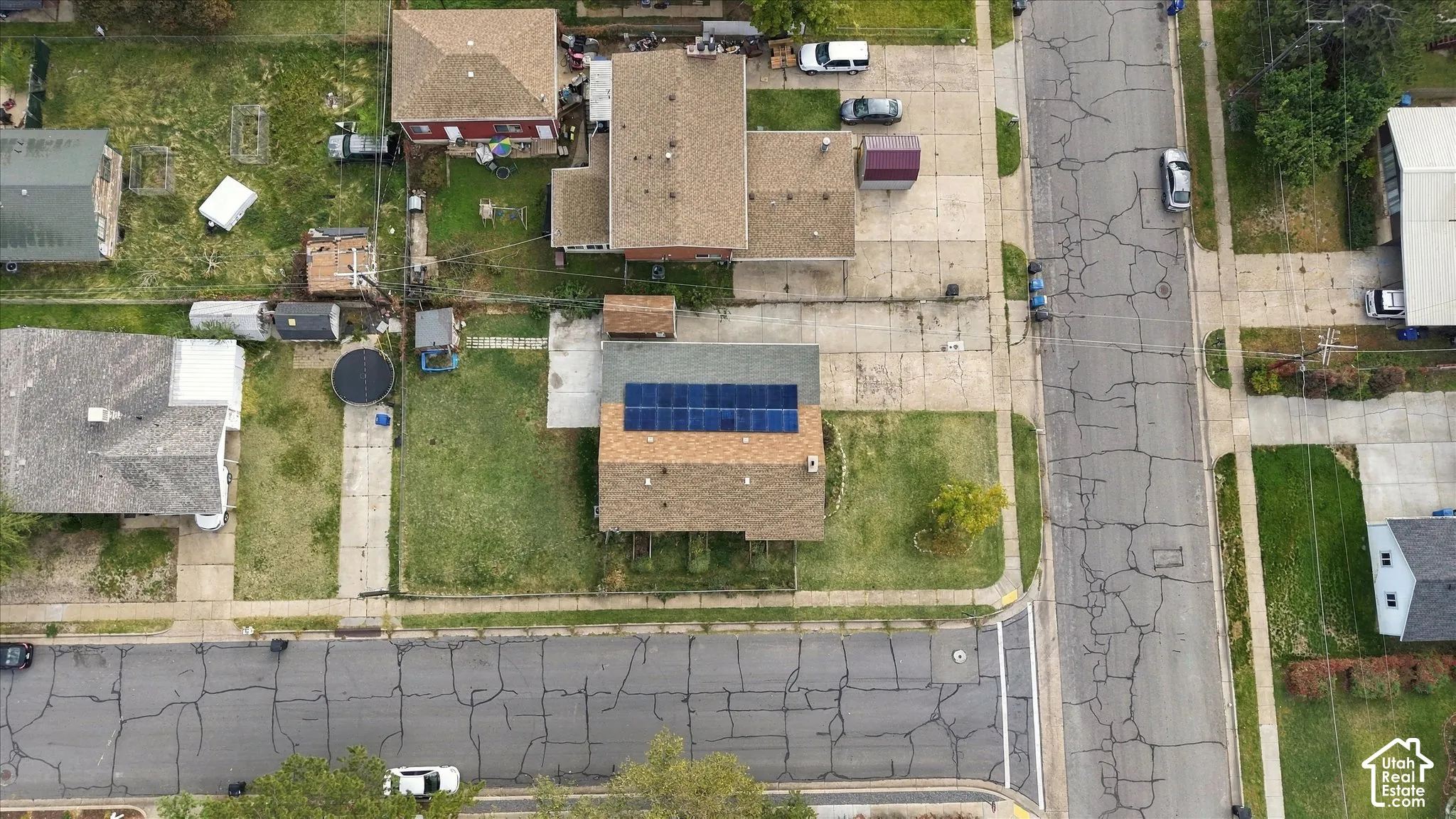 Aerial view of a residential lot featuring a multi-level asphalt shingle roof equipped with a grid of solar panels, a concrete driveway, and a fenced-in grassy yard
