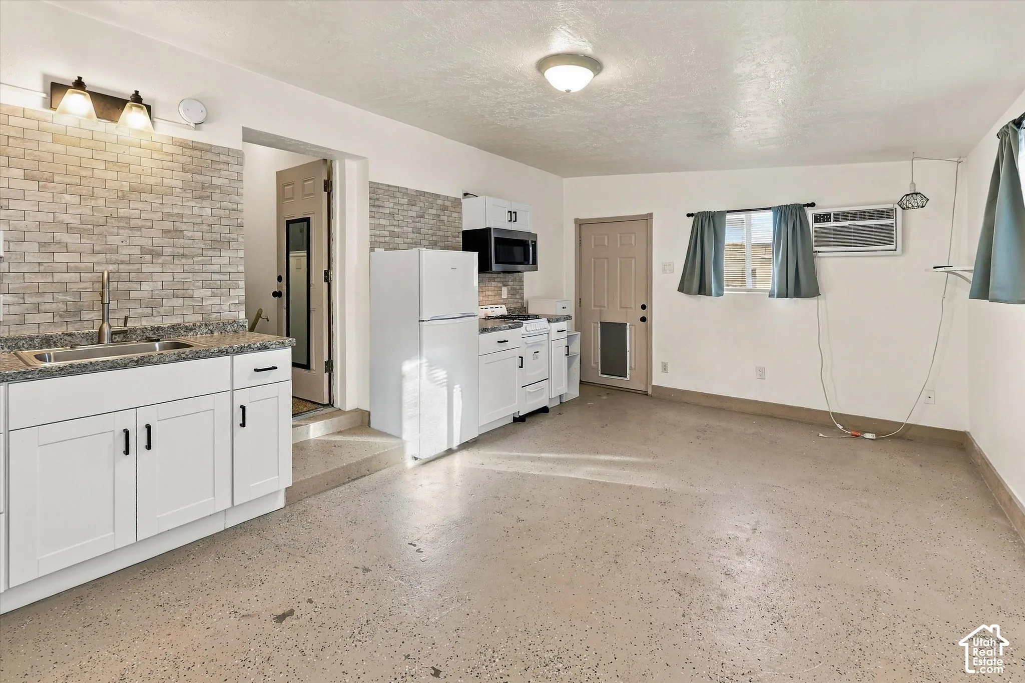 Kitchen area featuring white cabinetry, stone-finish countertops, brick-style tile backsplash, stainless steel sink with pull-down faucet, and overhead microwave.  This room can also be used as a separate dwelling unit.