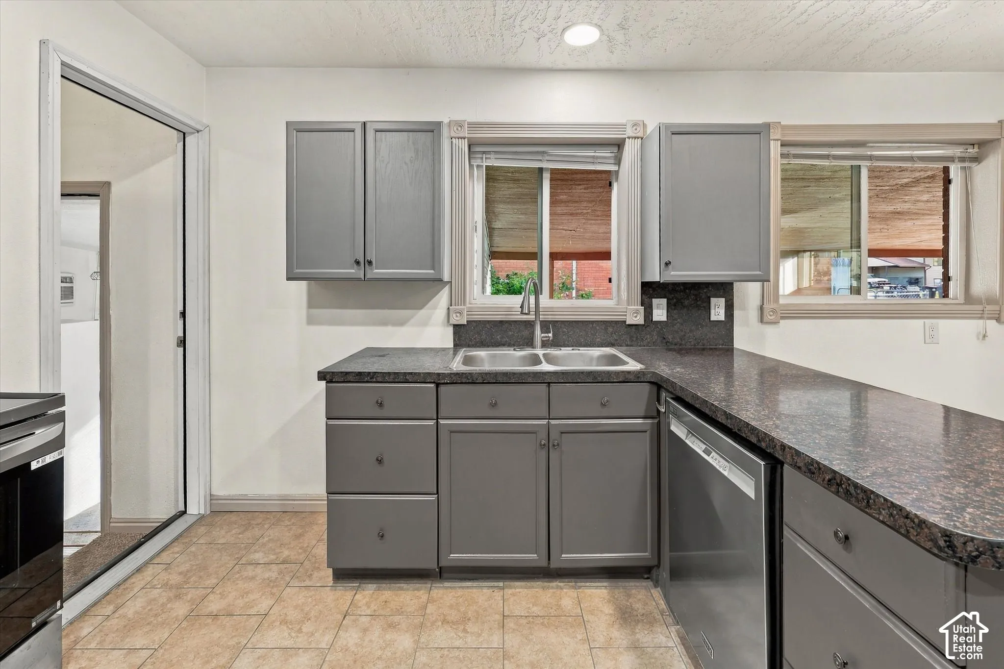 Kitchen area featuring grey cabinetry, dark stone-finish countertops, a dual-basin stainless steel sink with a high-arch faucet, a stainless steel dishwasher, and ceramic tile flooring