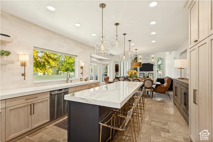 Kitchen featuring a large central island with a stone-finish countertop, light wood cabinetry, stone-finish tile flooring, textured stone wall accents, and glass pendant lighting