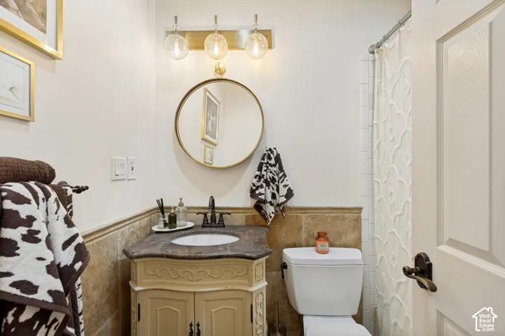 Bathroom featuring a vanity with a dark stone countertop, an undermount sink, a circular gold-rimmed mirror, three-bulb vanity lighting, and stone wainscoting