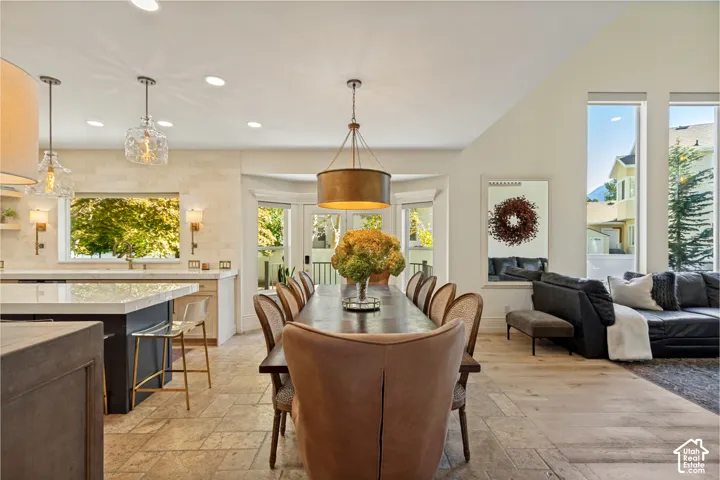 Open-concept space featuring vaulted ceilings, oversized windows, stone-finish flooring, a kitchen island with pendant lighting, and textured stone backsplash