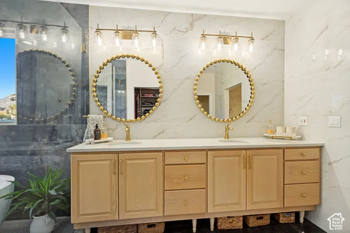 Dual vanity featuring light wood-finish cabinetry, quartz countertops, circular gold-tone framed mirrors, and gold-tone wall-mounted lighting fixtures against a full-height tile backsplash
