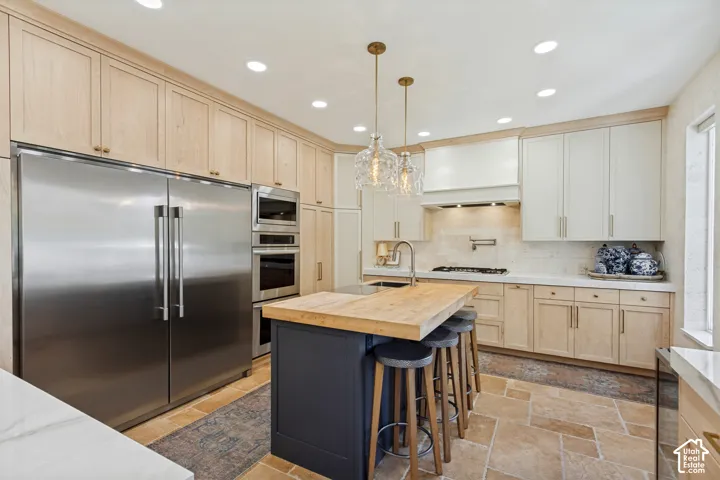 Kitchen featuring light wood-finish cabinetry, stainless steel double wall ovens, a center island with a wood countertop, stone tile flooring, and dual glass pendant lights