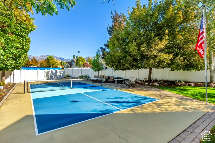 Backyard pickleball court featuring a multi-toned blue playing surface, white perimeter fencing, manicured grass, mature trees, and a flag pole