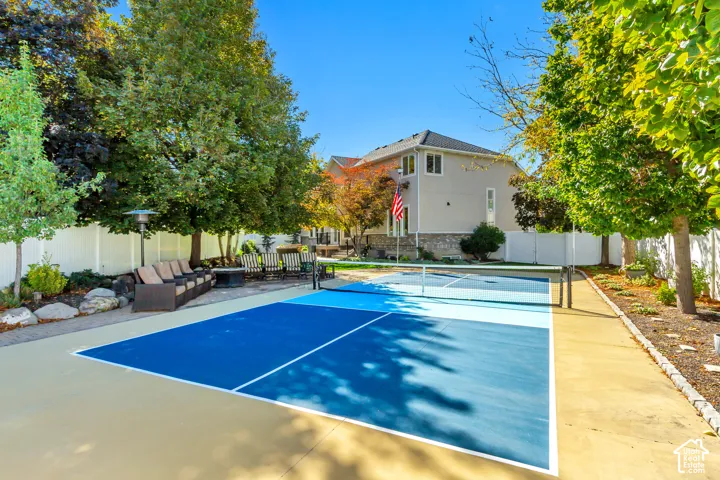 Private sports court featuring a multi-toned blue surface, enclosed by a white vinyl privacy fence, stone-bordered landscaping, and mature trees