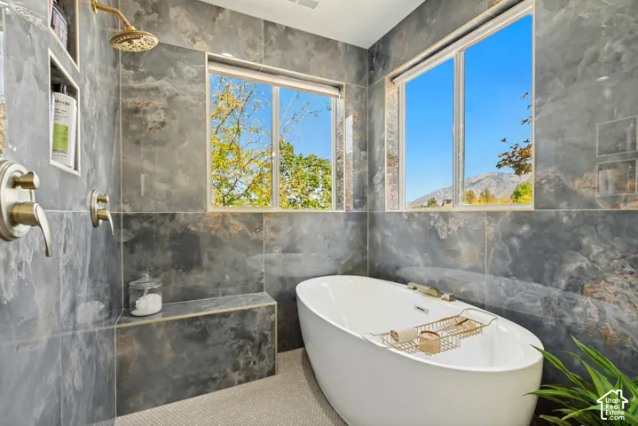 Freestanding soaking tub with a deck-mounted faucet, floor-to-ceiling stone-tiled walls, built-in recessed wall shelving, and dual windows with mountain scenery