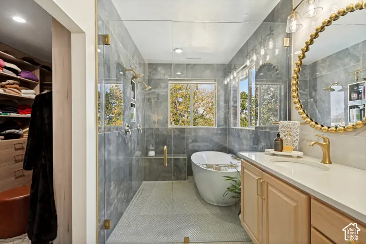 Bathroom featuring stone-tile walls, a glass-enclosed shower with dual showerheads, a freestanding soaking tub, light wood cabinetry with a quartz countertop, and gold-tone fixtures