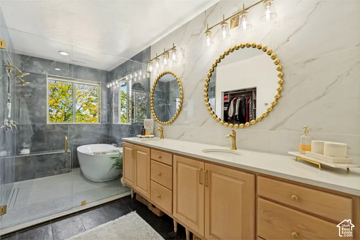 Dual-vanity cabinetry with a light-finish wood tone, solid-surface countertop, gold-toned hardware, large-format veined wall tile, and a glass-enclosed wet room featuring a freestanding soaking tub