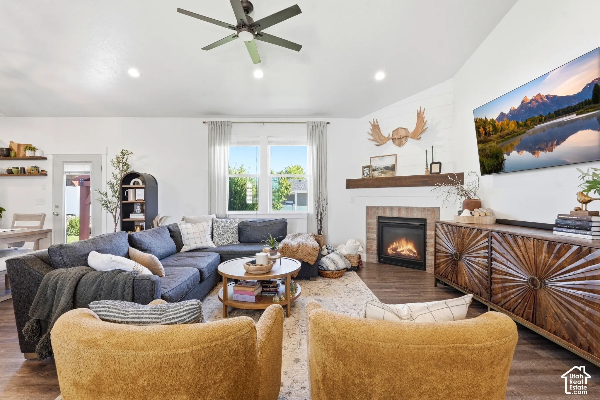Vaulted ceiling with a central ceiling fan, fireplace featuring stone-surround, wood-finish mantel, horizontal shiplap detailing, and wood-finish flooring
