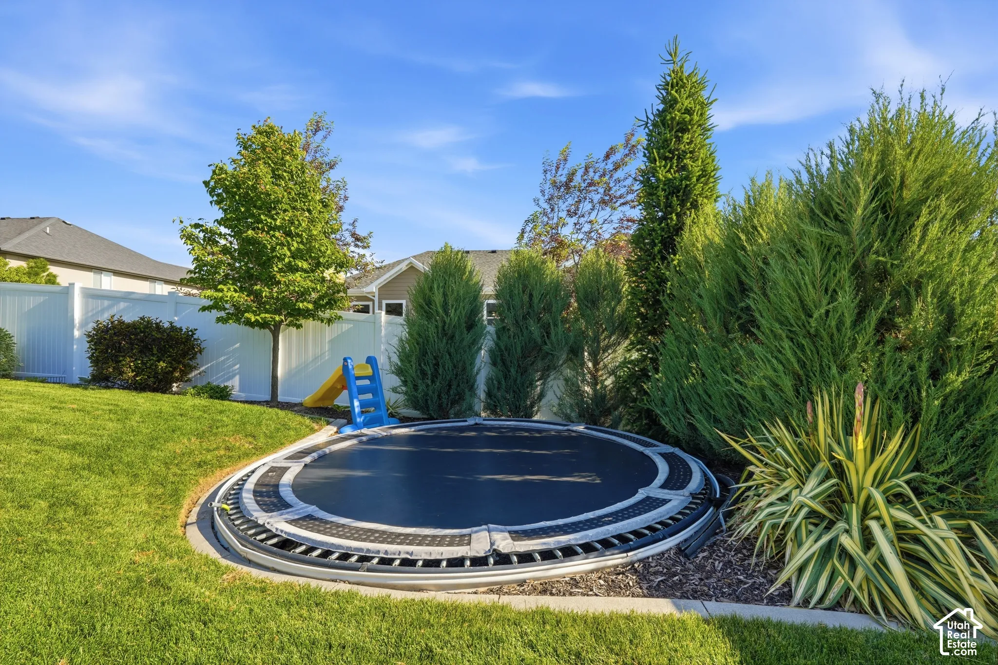 Backyard featuring a sunken in-ground trampoline, white vinyl perimeter fencing, stone edging, mature evergreen landscaping, and a manicured lawn