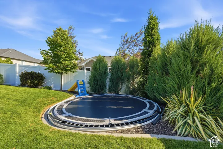 Backyard featuring a sunken in-ground trampoline, white vinyl perimeter fencing, stone edging, mature evergreen landscaping, and a manicured lawn