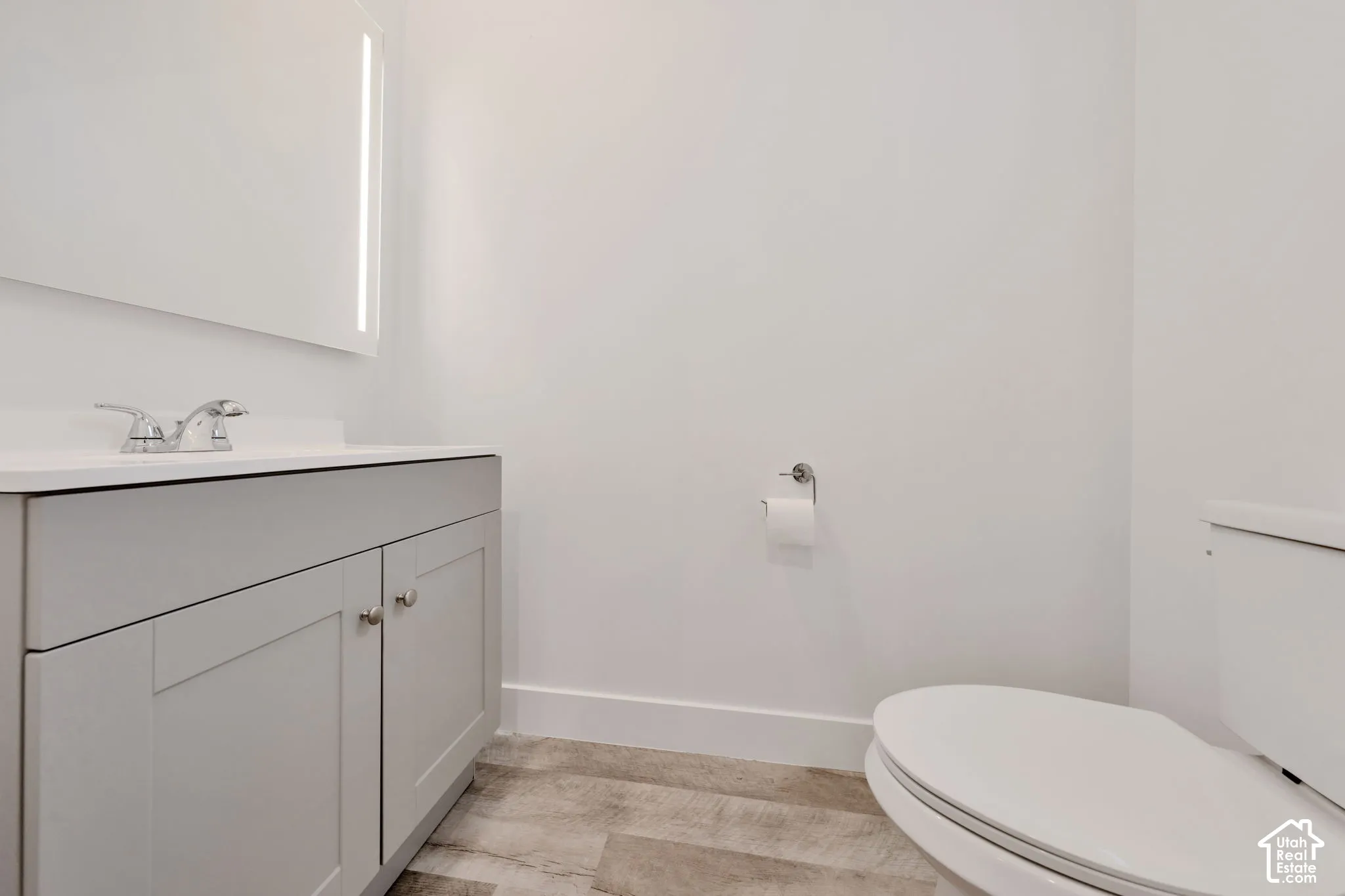 Grey shaker-style vanity with a solid surface countertop, chrome dual-handle faucet, illuminated rectangular wall mirror, and wood-finish flooring