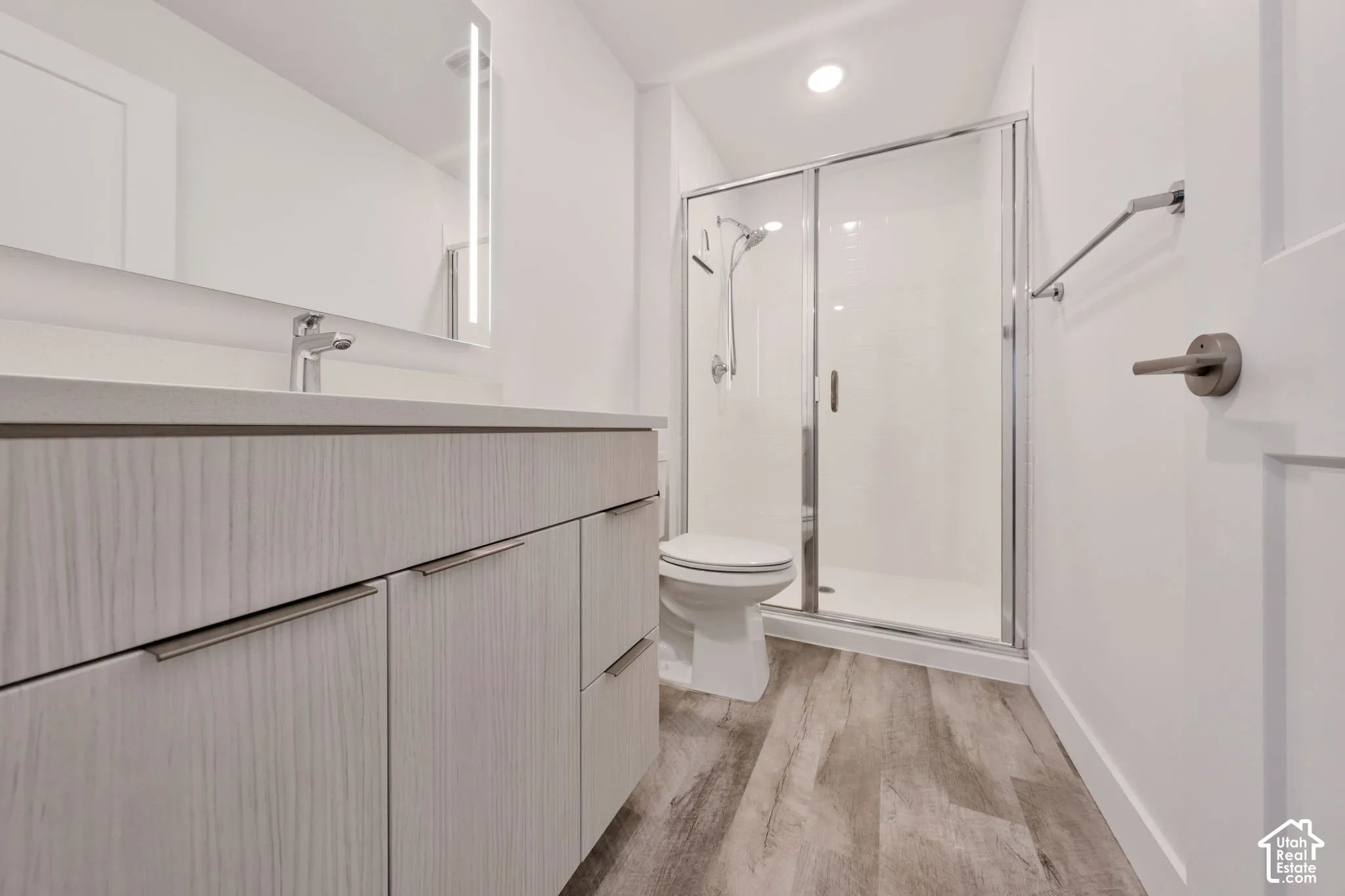 Bathroom featuring wood-finish cabinetry, light-toned countertop with integrated sink, backlit vanity mirror, wood-finish flooring, and glass-enclosed shower