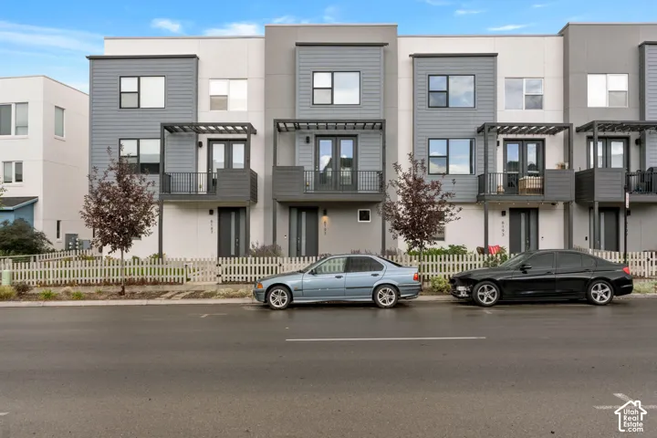Modern multi-story exterior featuring fiber cement siding, metal balcony pergolas, wrought iron railings, and private white picket fencing