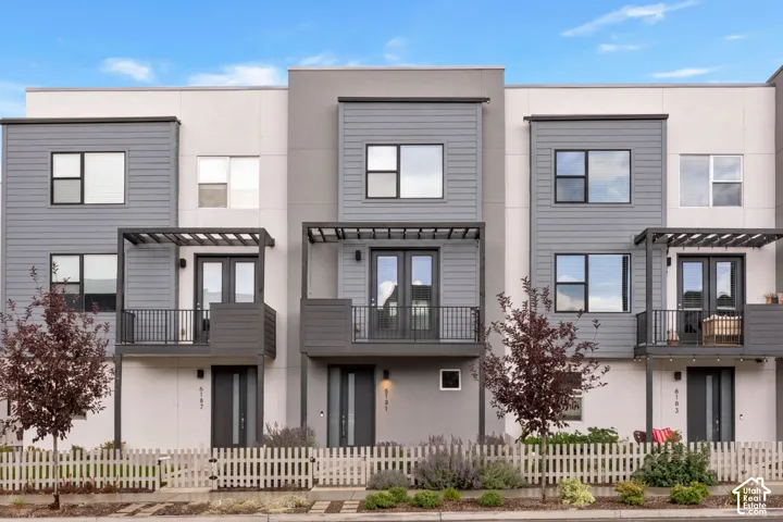 Modern multi-story exterior featuring a gray horizontal siding and smooth stucco facade, metal-framed balconies, pergola-style overhead structures, and metal picket railings