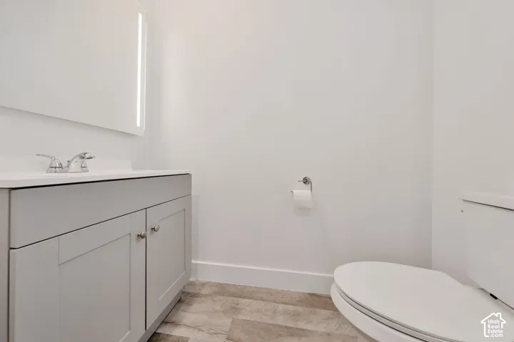 Grey shaker-style vanity with a solid surface countertop, chrome dual-handle faucet, illuminated rectangular wall mirror, and wood-finish flooring