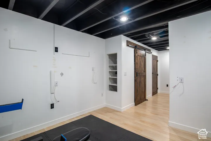 Open-concept interior featuring light wood-finish flooring, exposed painted ceiling joists, recessed lighting, integrated wall shelving, and a rustic wood sliding door with black hardware