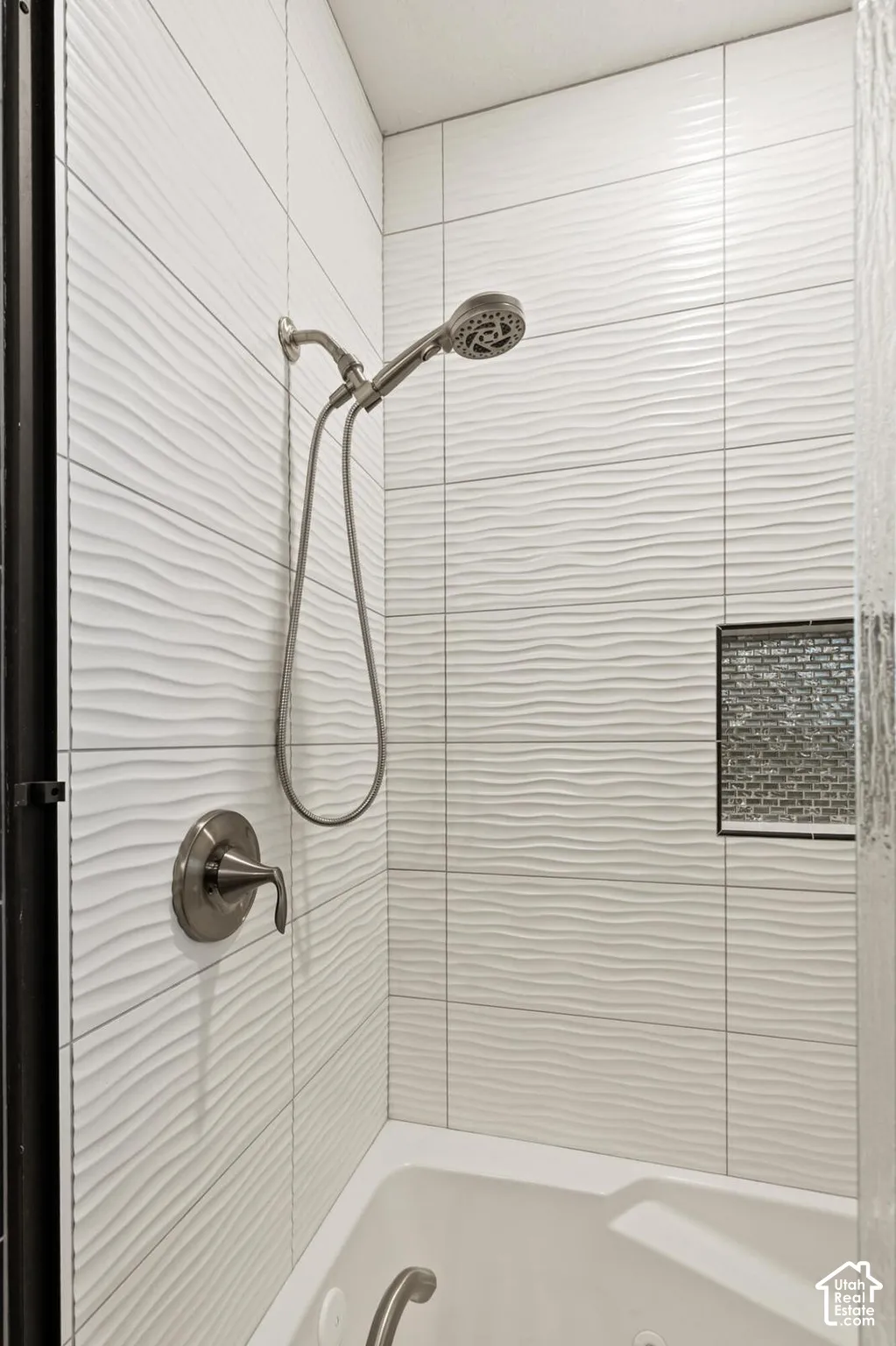 Shower area featuring textured white wall tile, brushed metallic handheld shower head with single-handle mixing valve, and a decorative tile-lined recessed wall niche