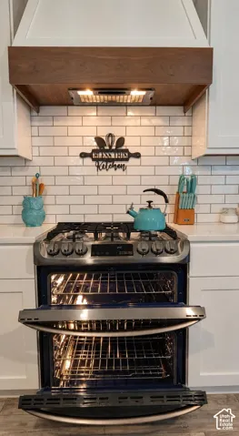 White shaker-style cabinetry, double-oven stainless steel gas range, subway tile backsplash, and custom wood-trim vent hood