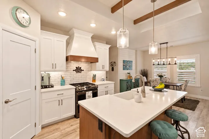 Kitchen featuring a central island with a quartz countertop and an undermount sink, custom white cabinetry, a wood-accented range hood, subway tile backsplash, and wood-finish flooring